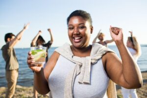 Smiling woman holding a refreshing drink at the beach, celebrating healthy lifestyle choices and enjoying a weight loss journey with supportive habits and positive energy.