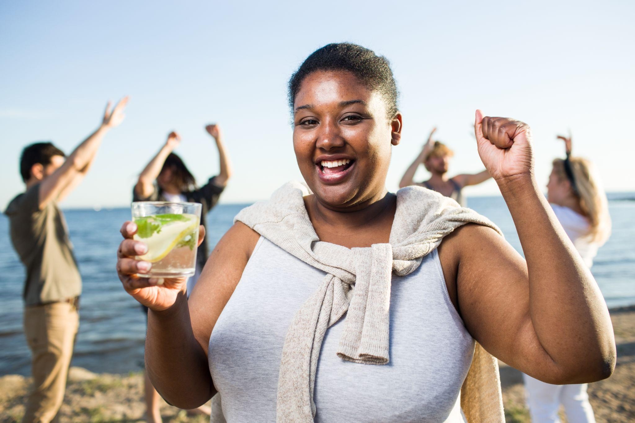 Smiling woman holding a refreshing drink at the beach, celebrating healthy lifestyle choices and enjoying a weight loss journey with supportive habits and positive energy.