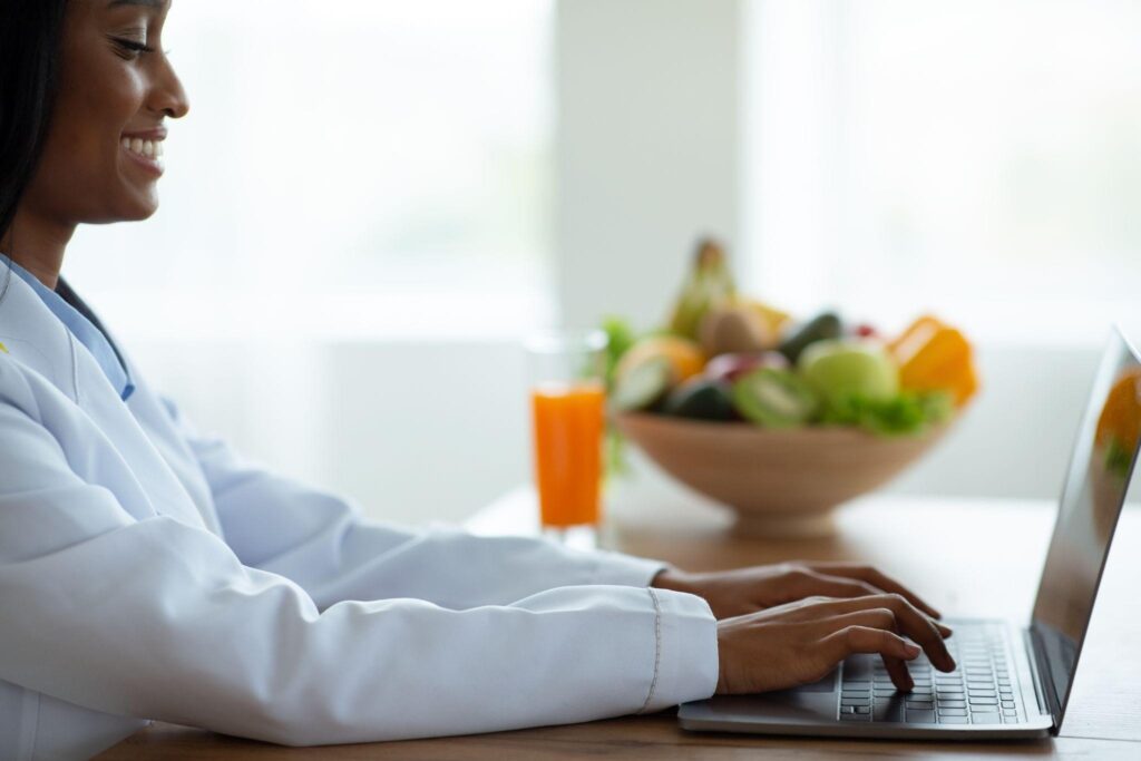Close-up of a virtual weight loss specialist typing on a laptop with healthy fruits in the background. Represents online metabolic reset coaching and natural nutrition support.