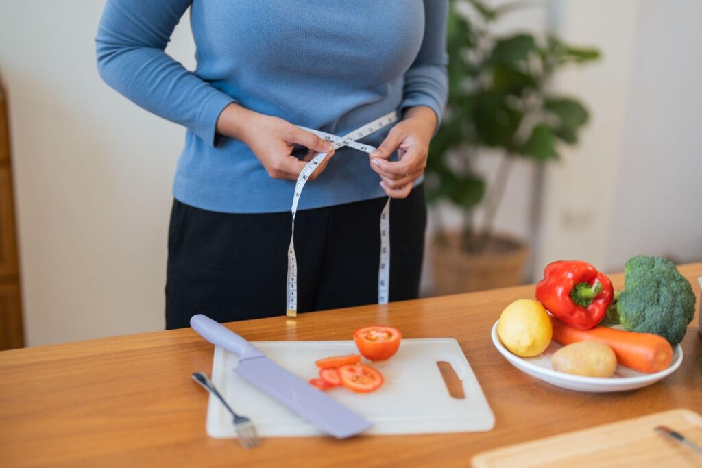 Asian woman measuring her waist at home while preparing healthy vegetables, representing success from weight loss programs in Eau Claire, WI, targeting inflammation.