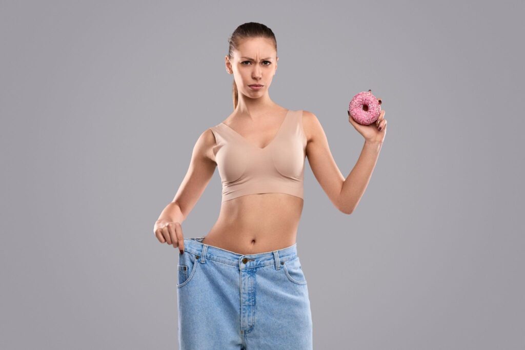 Young woman holding a donut while showing loose jeans, representing results after following a metabolic reset method for sustainable weight loss in Wisconsin.
