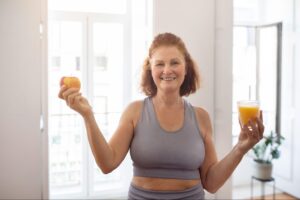 A smiling older woman holding an apple and a glass of juice, representing healthy habits that support metabolism, as adults wonder when their metabolism slows down.