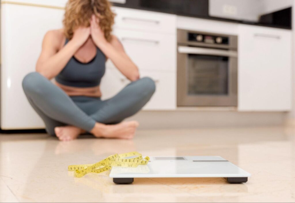 A woman sitting on her kitchen floor looking frustrated near a scale, illustrating slow metabolism symptoms and the challenges adults face long before they ask when does your metabolism slow down.